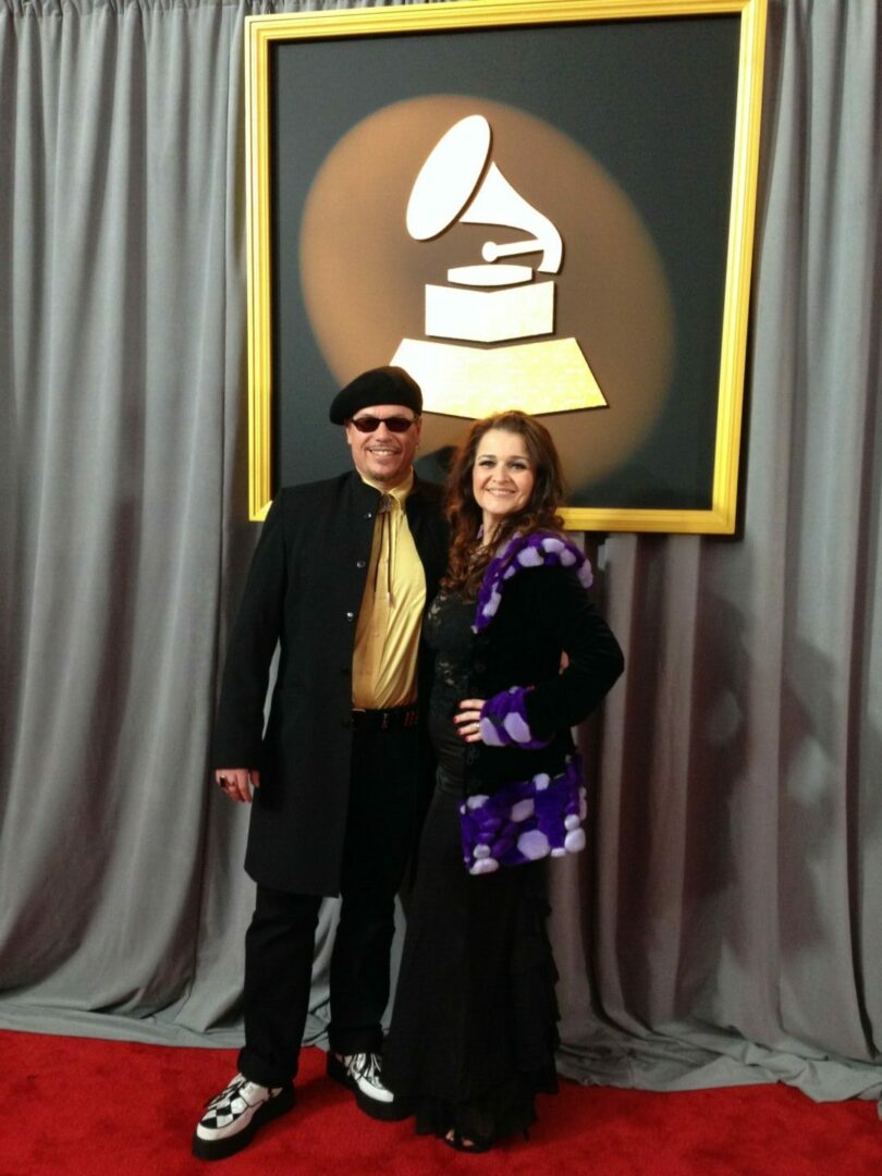 Two people standing in front of a Grammy Award logo on a black background, with gray curtains and a red carpet.