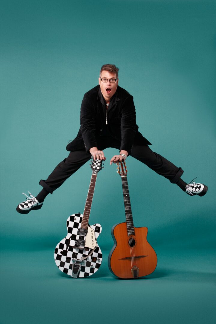 Person jumping with legs spread, holding two guitars, one checkered and one wooden, against a teal background.