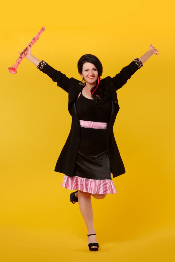 Woman in black dress with pink accents, smiling, holding a pink toy, arms raised, standing on one leg against yellow background.