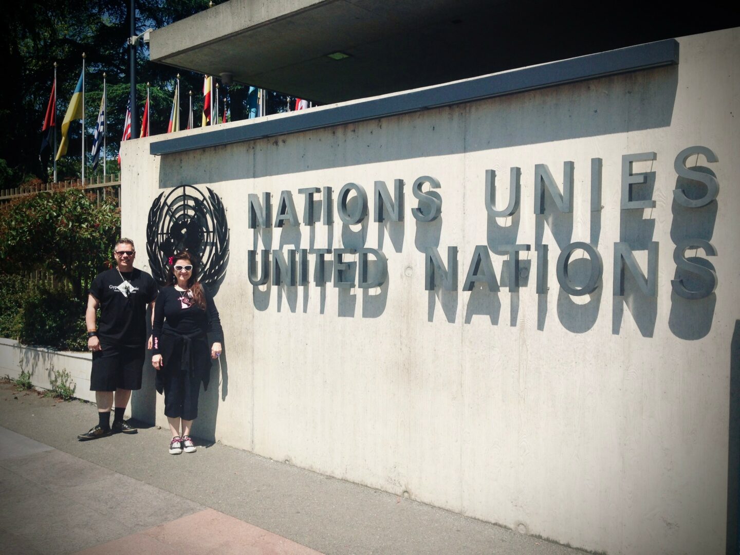 Two people stand in front of a United Nations sign on a concrete wall with flags in the background.