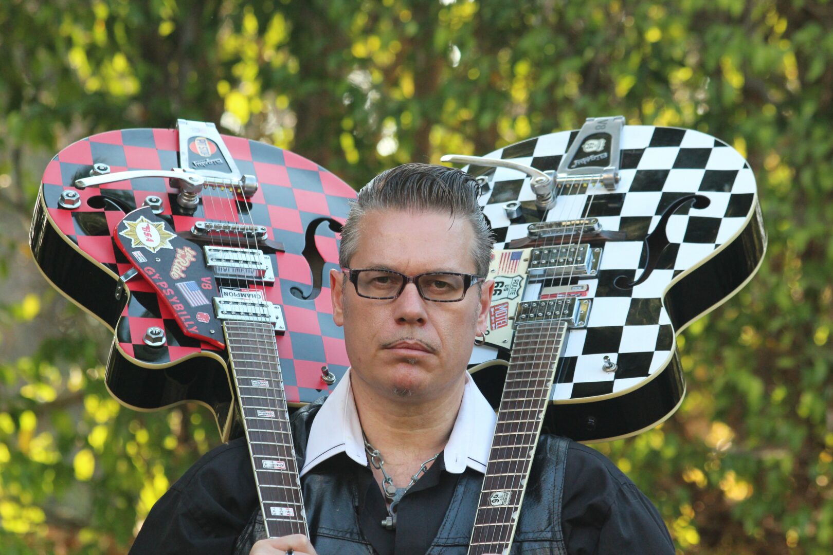 Person with glasses and white shirt under black jacket, holding two guitars with checkered patterns, outdoors with green foliage.