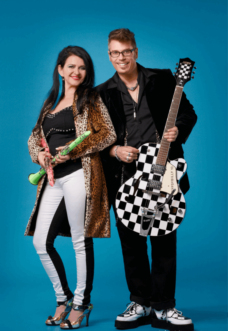 A man and woman stand together against a blue background, holding musical instruments and smiling.