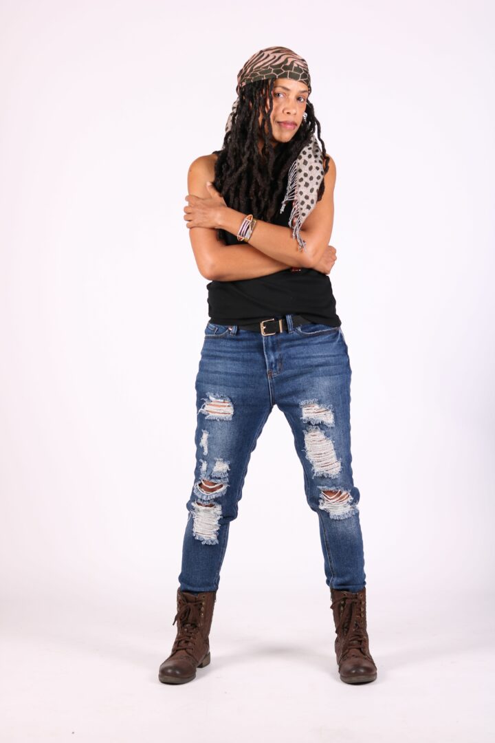 Young woman with long curly hair wearing a headband, sleeveless top, ripped jeans, and boots, standing with arms crossed.