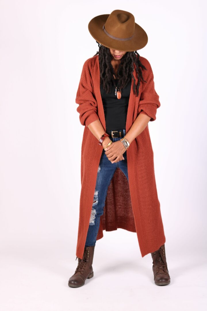 Woman wearing a large brown hat, long red coat, black top, ripped jeans, and brown boots, standing with hands clasped.