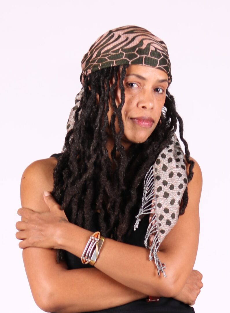 Woman with long dreadlocks wearing a patterned headscarf and scarf, crossing arms, looking at camera.