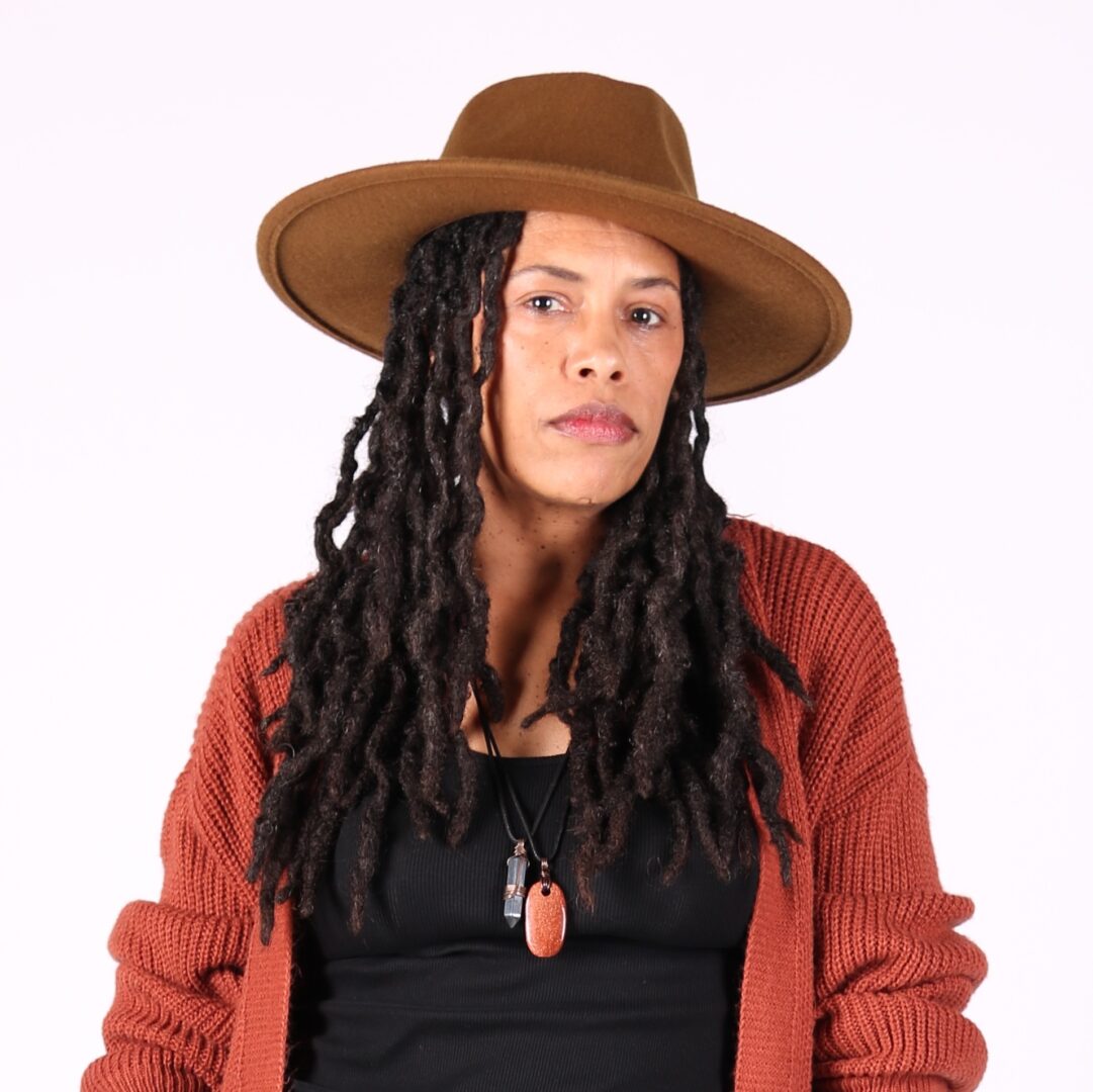 Woman with long curly hair wearing a wide-brimmed hat, red cardigan, and black top, looking at the camera.