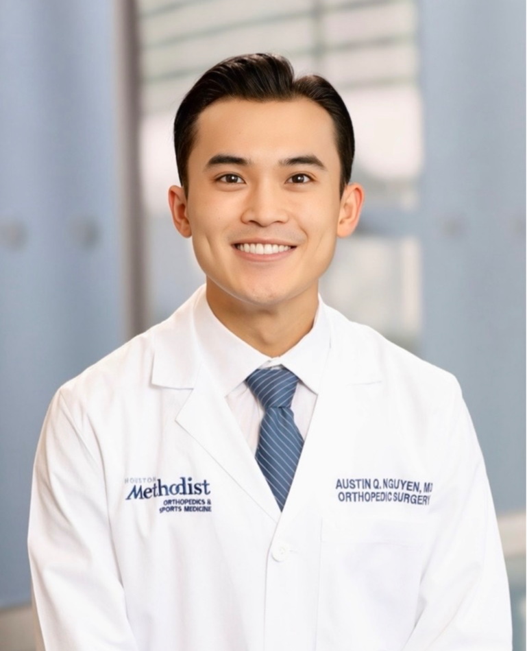 Young man in white medical coat smiling, standing indoors with blurred background.