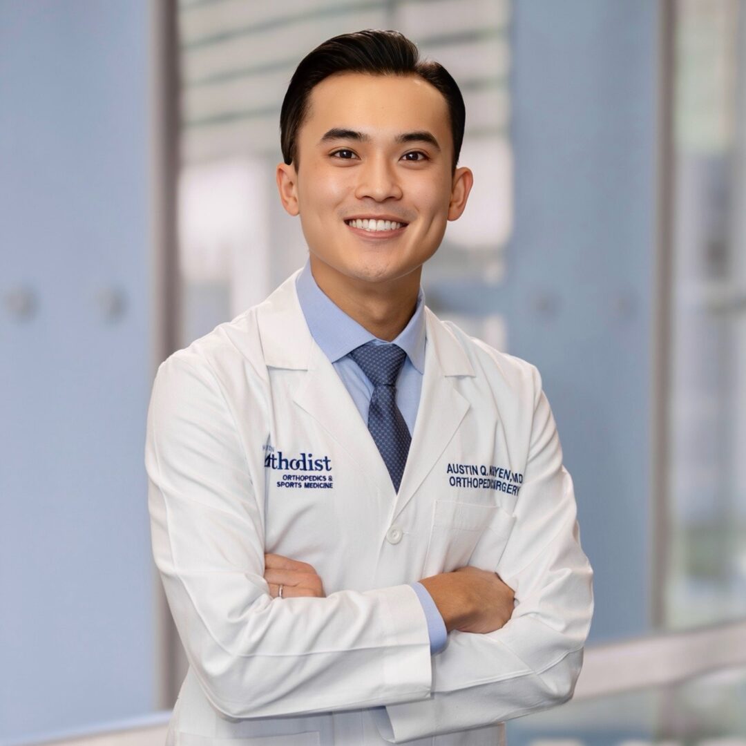 Young male doctor in white coat smiling with arms crossed in hospital setting.
