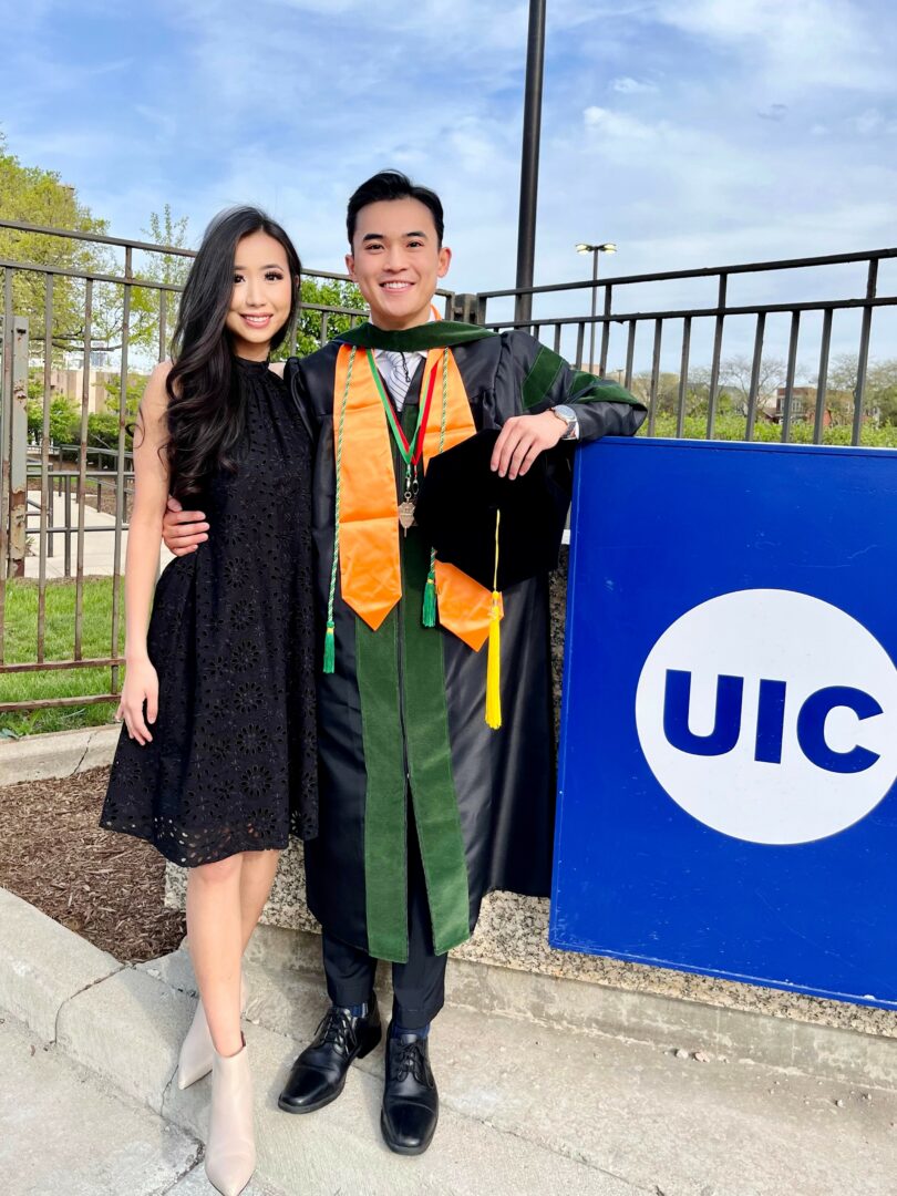 Graduate in cap and gown with a woman in a black dress outdoors near a UIC sign.