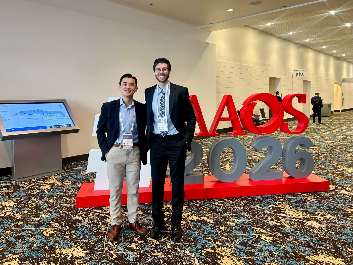 Two men in suits standing in front of large red and gray 'KAOS 2026' sign in conference hall.