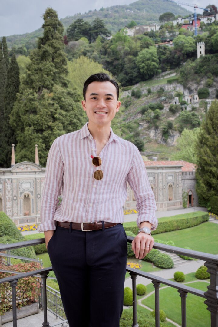Young man in striped shirt and dark pants standing outdoors with greenery and historic architecture in background.