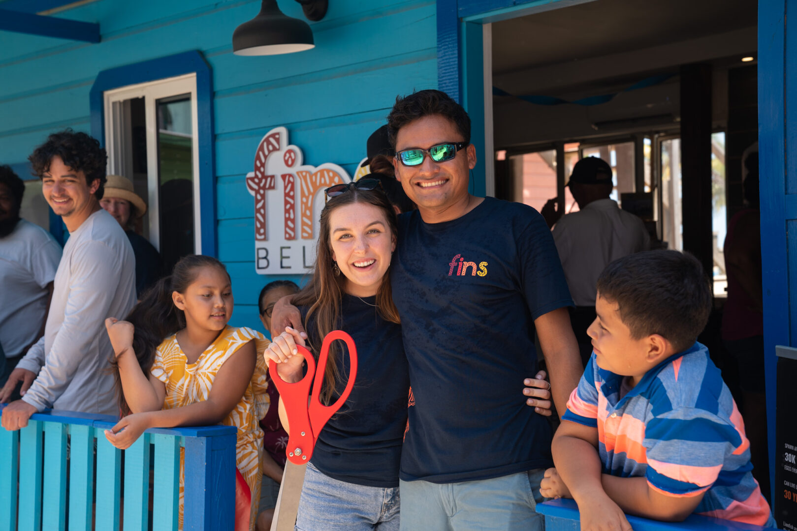 Two smiling people, a woman and a man, stand outside a blue building with children nearby, one girl and one boy.