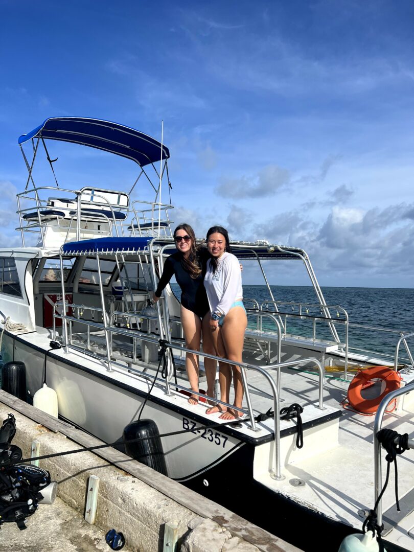 Two women stand on a boat docked at a marina, smiling, with water and sky in the background.