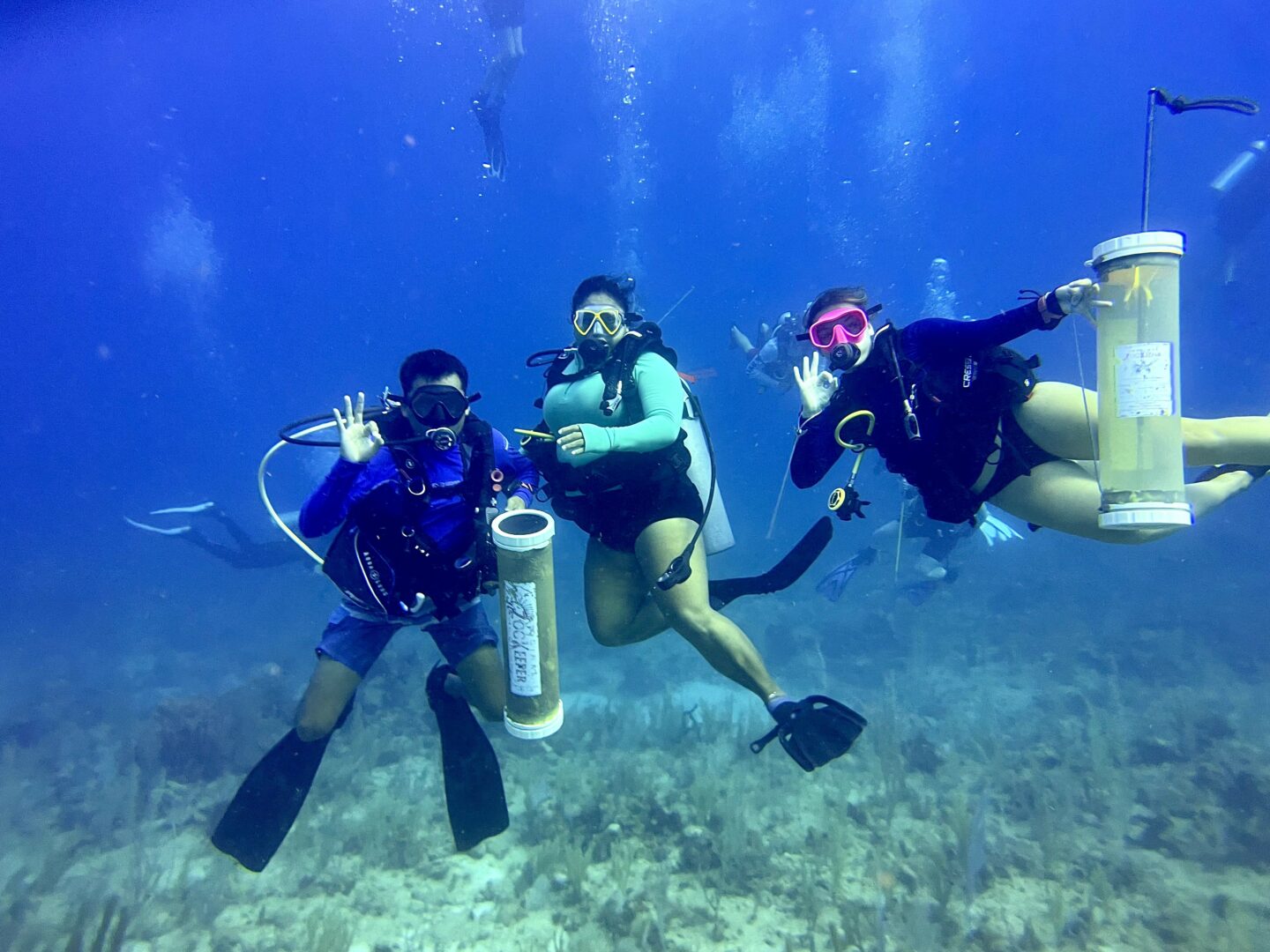 Three scuba divers underwater, holding tanks, making peace signs, with a blue background and sea floor visible.
