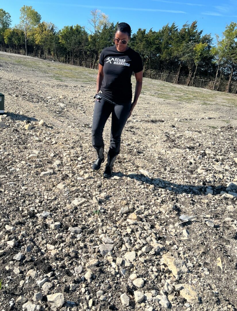 Person walking on rocky ground outdoors, wearing sunglasses, black t-shirt, and black pants, with trees and blue sky in background.