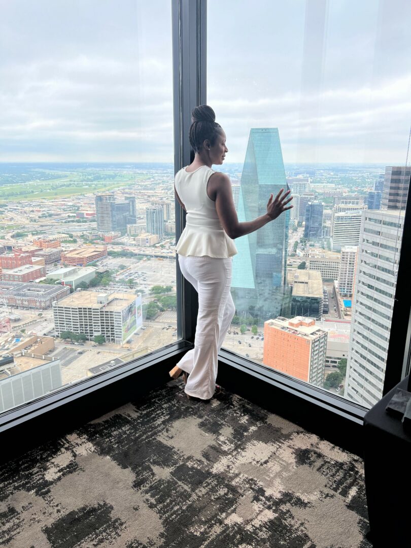 Woman standing by window with cityscape view, touching glass with one hand, wearing white outfit, hair in bun.