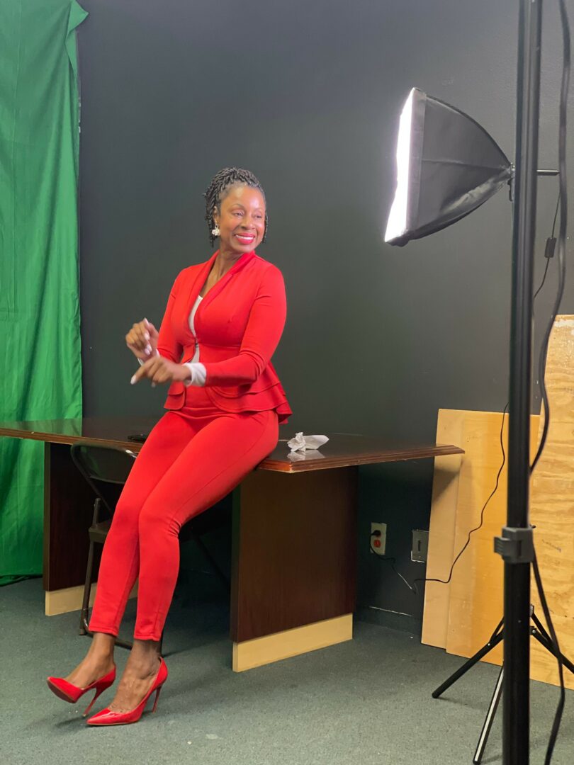 Woman in red suit and heels sitting on desk, smiling, in a studio with green screen and lighting equipment.