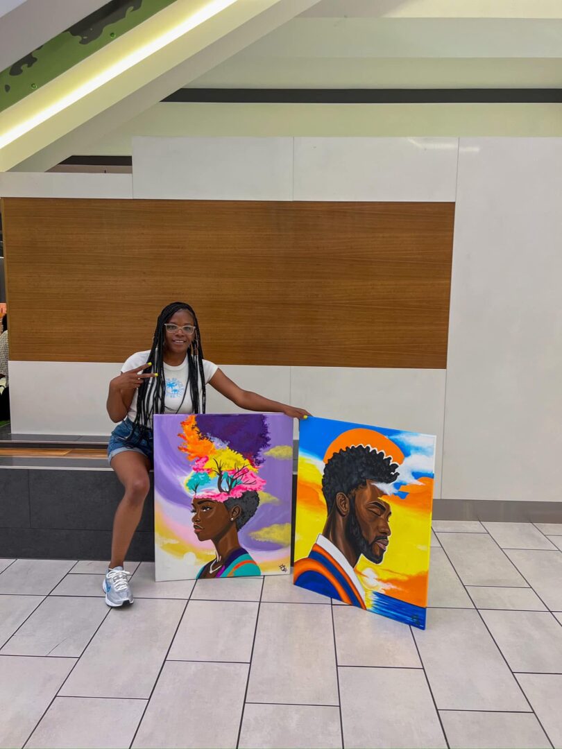 Person with long braids sitting next to two colorful portraits on the floor in a modern indoor space.