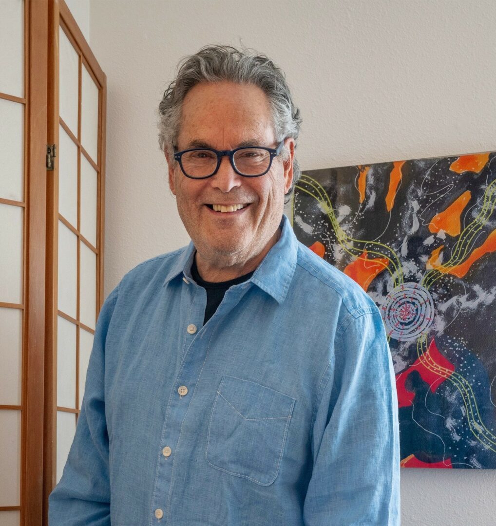 Smiling man with glasses and gray hair wearing a blue shirt indoors.