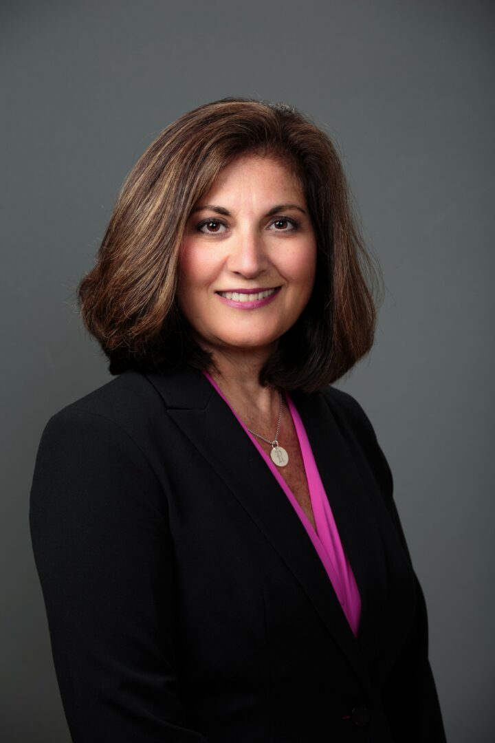 Woman with shoulder-length brown hair, smiling, wearing a black blazer and pink blouse, against gray background.