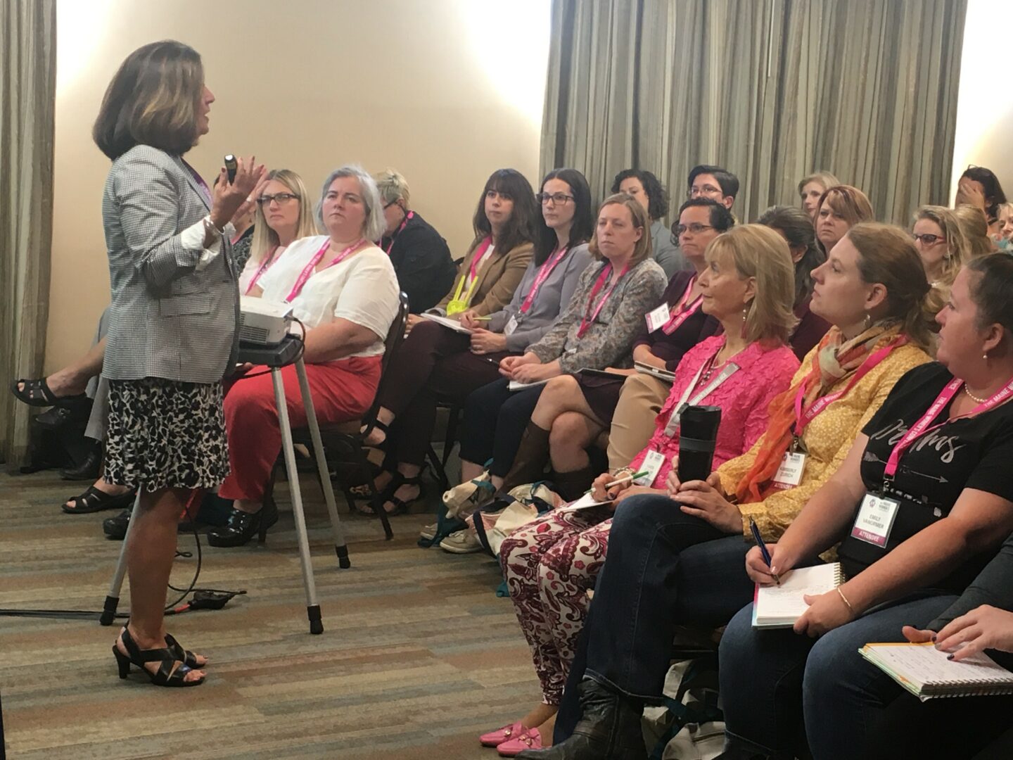 A woman stands and speaks to a seated audience in a conference room with curtains and wooden floor.