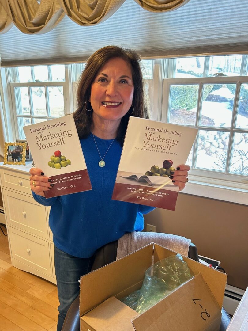 Woman holding two books titled 'Marketing Yourself' in a room with large windows and a cardboard box on a table.