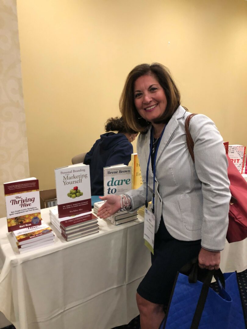 Woman smiling at a book display table with books and promotional materials, wearing a white blazer and dark skirt.
