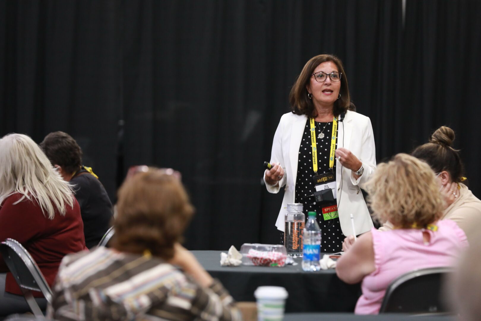 Woman speaking to group in a room with black curtains, wearing glasses, white blazer, and black shirt with yellow dots.