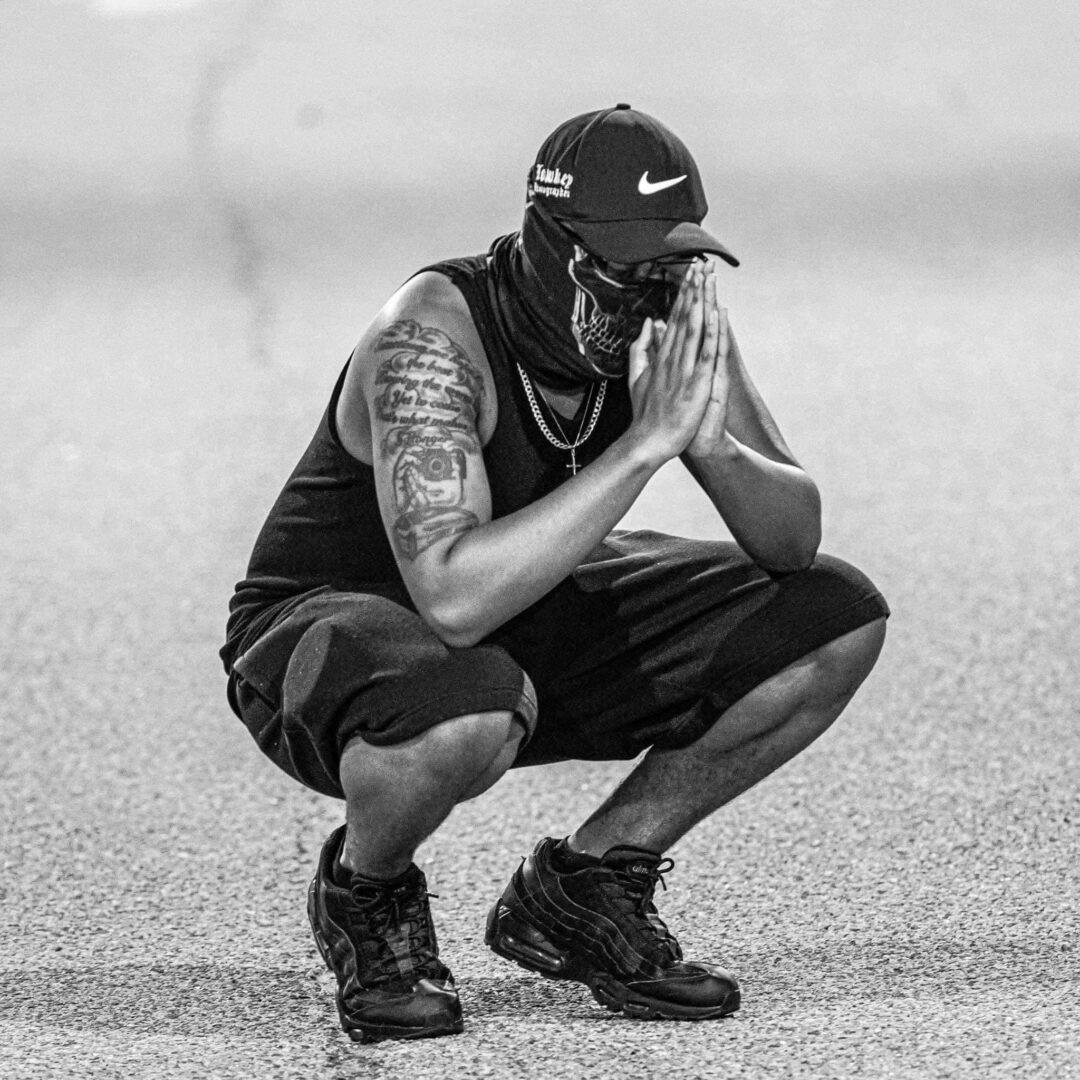 Person squatting outdoors, wearing a cap, sleeveless shirt, and sneakers, with hands pressed together in front of face.