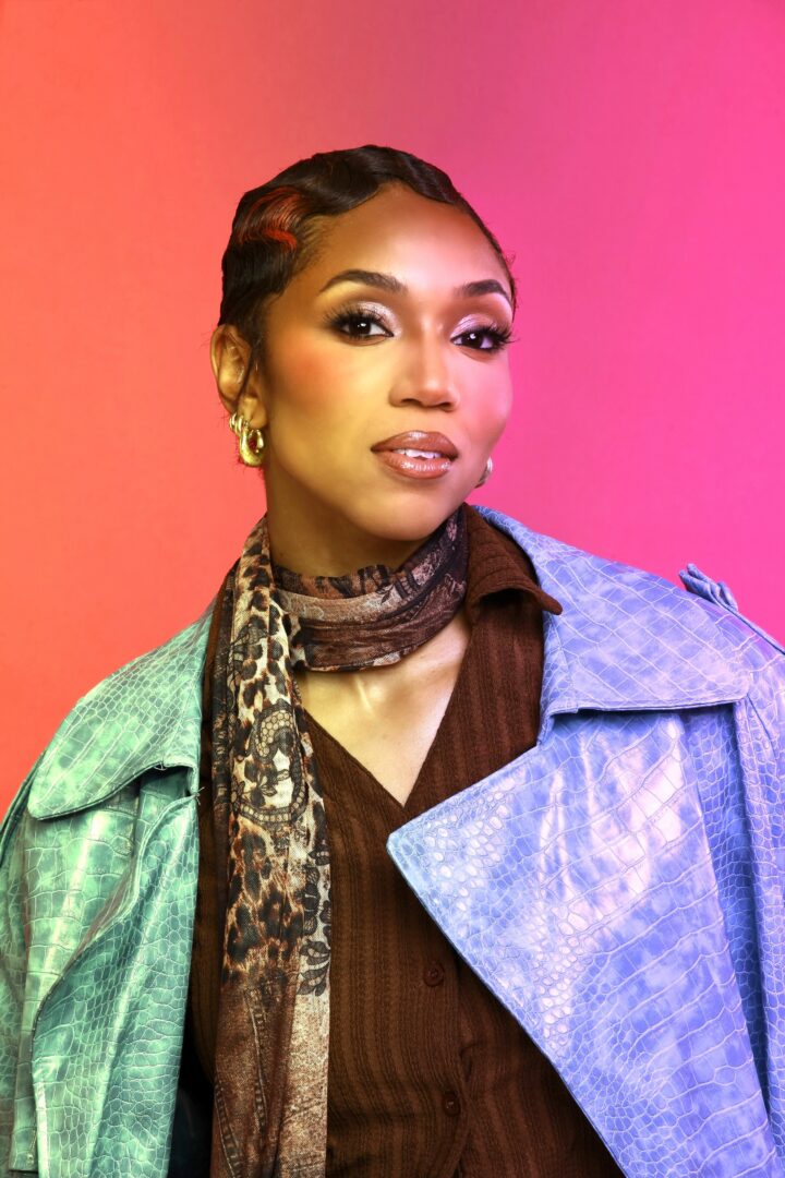 Woman with braided hair, wearing gold earrings, a patterned scarf, and a blue jacket against a pink and orange background.