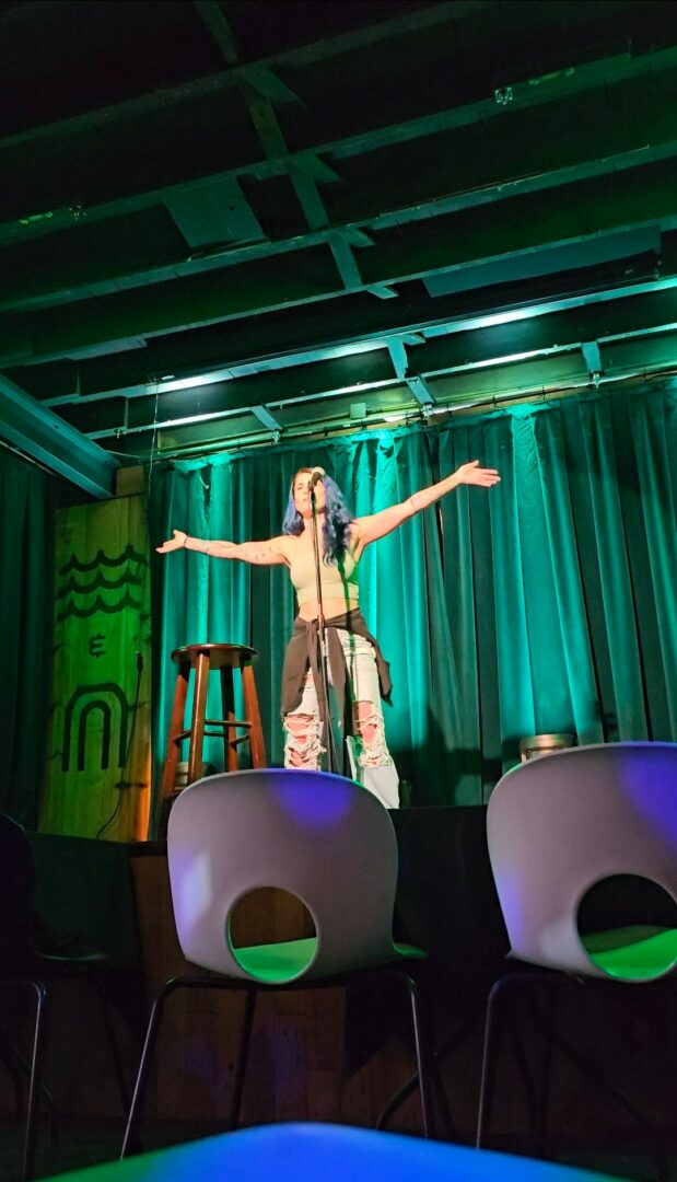 Performer on stage with arms outstretched, wearing a costume, in front of a green curtain and stage lights.