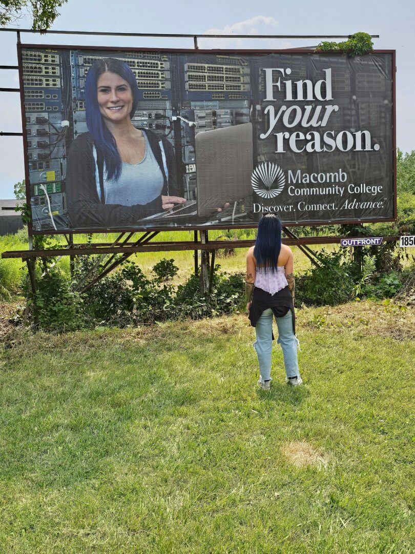 Person standing on grass near a large billboard with a woman's photo and text.