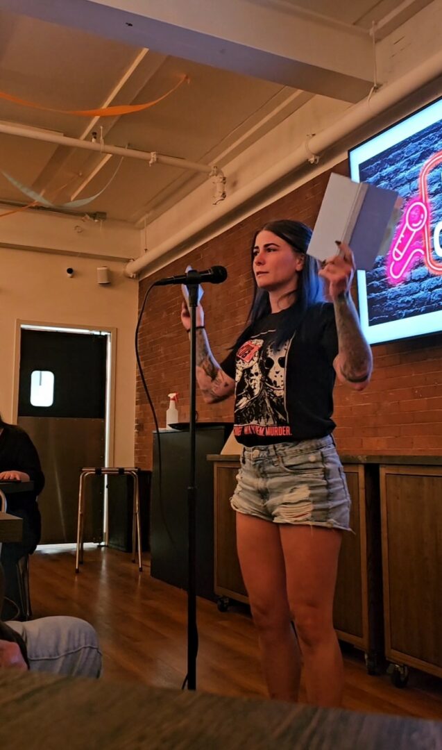 Young woman with dark hair, wearing a black graphic t-shirt and denim shorts, speaking into a microphone in a room.