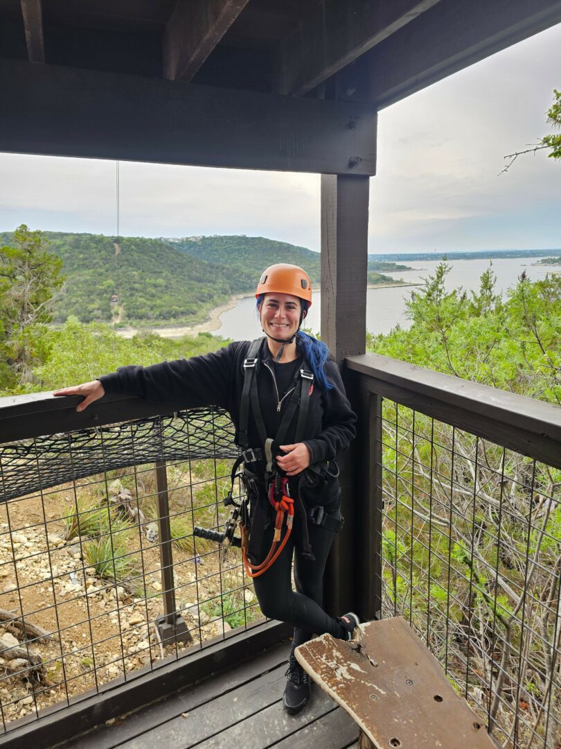 Person wearing a helmet and jacket standing on a balcony with a view of green landscape and sky.