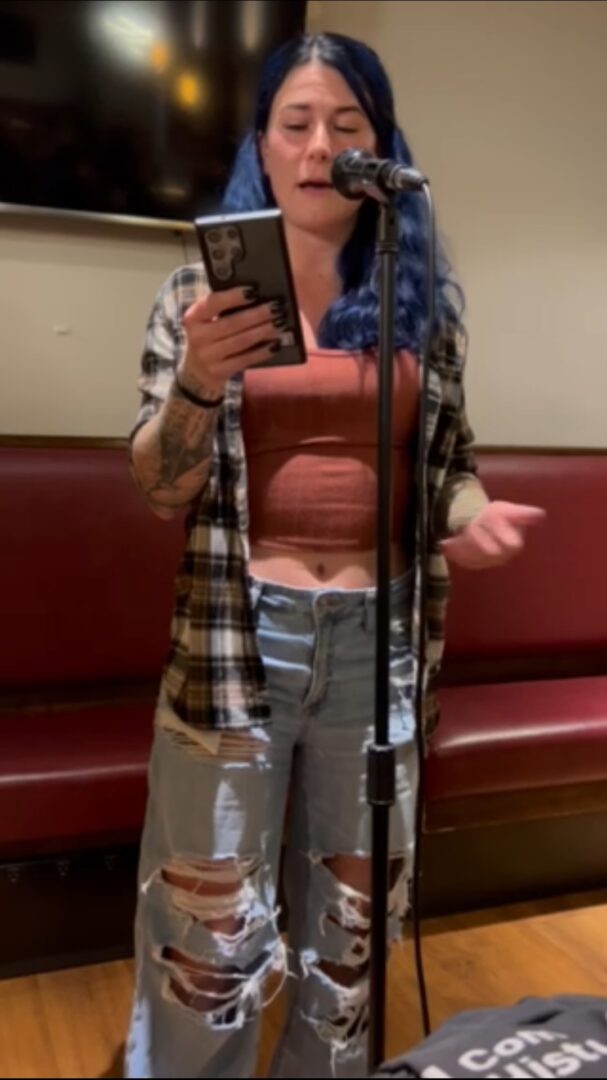 Young woman with blue hair reading from a phone into a microphone in a room with a red bench.