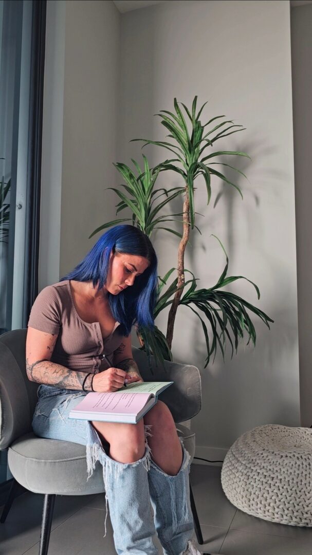 Woman with blue hair sitting on a chair, writing in a notebook, with a large plant behind her.