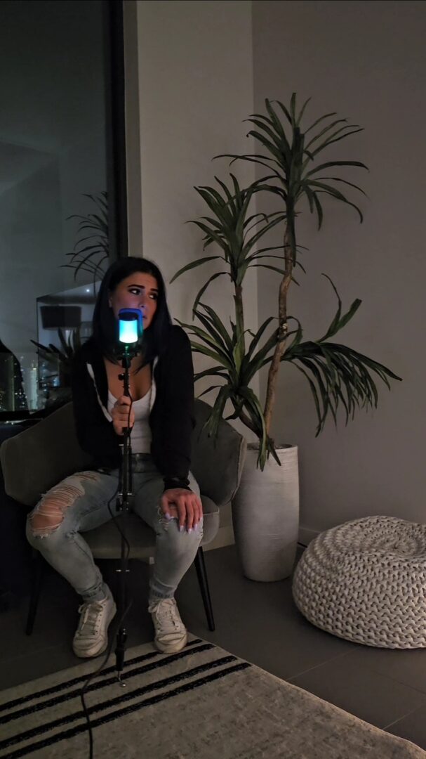 Person sitting on a chair with a mask, holding a phone, next to a large potted plant in a room.