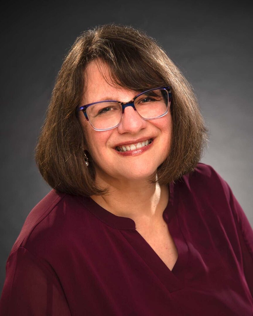 Smiling woman with shoulder-length brown hair, glasses, wearing a maroon top, against a dark background.