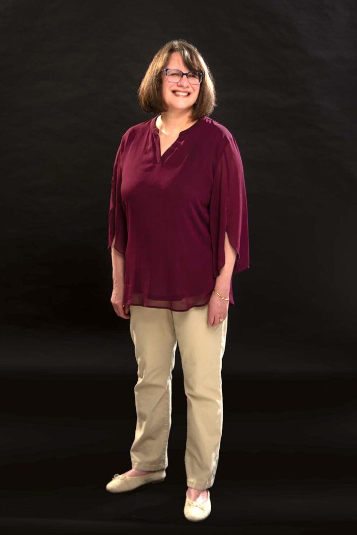 Woman with shoulder-length hair and glasses smiling, wearing a maroon blouse and beige pants, standing against a black background.