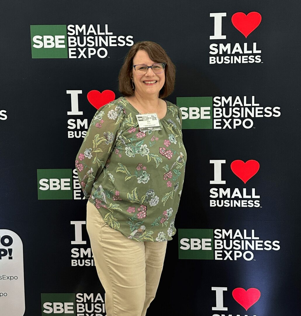 Woman with glasses smiling, wearing a green floral blouse and beige pants, standing in front of a black backdrop with logos.