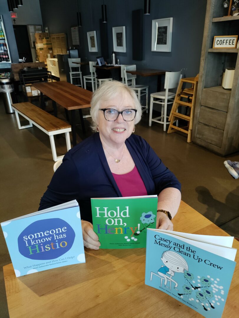 Woman with glasses and short blonde hair sitting at a table in a cafe, smiling, with three children's books displayed in front.
