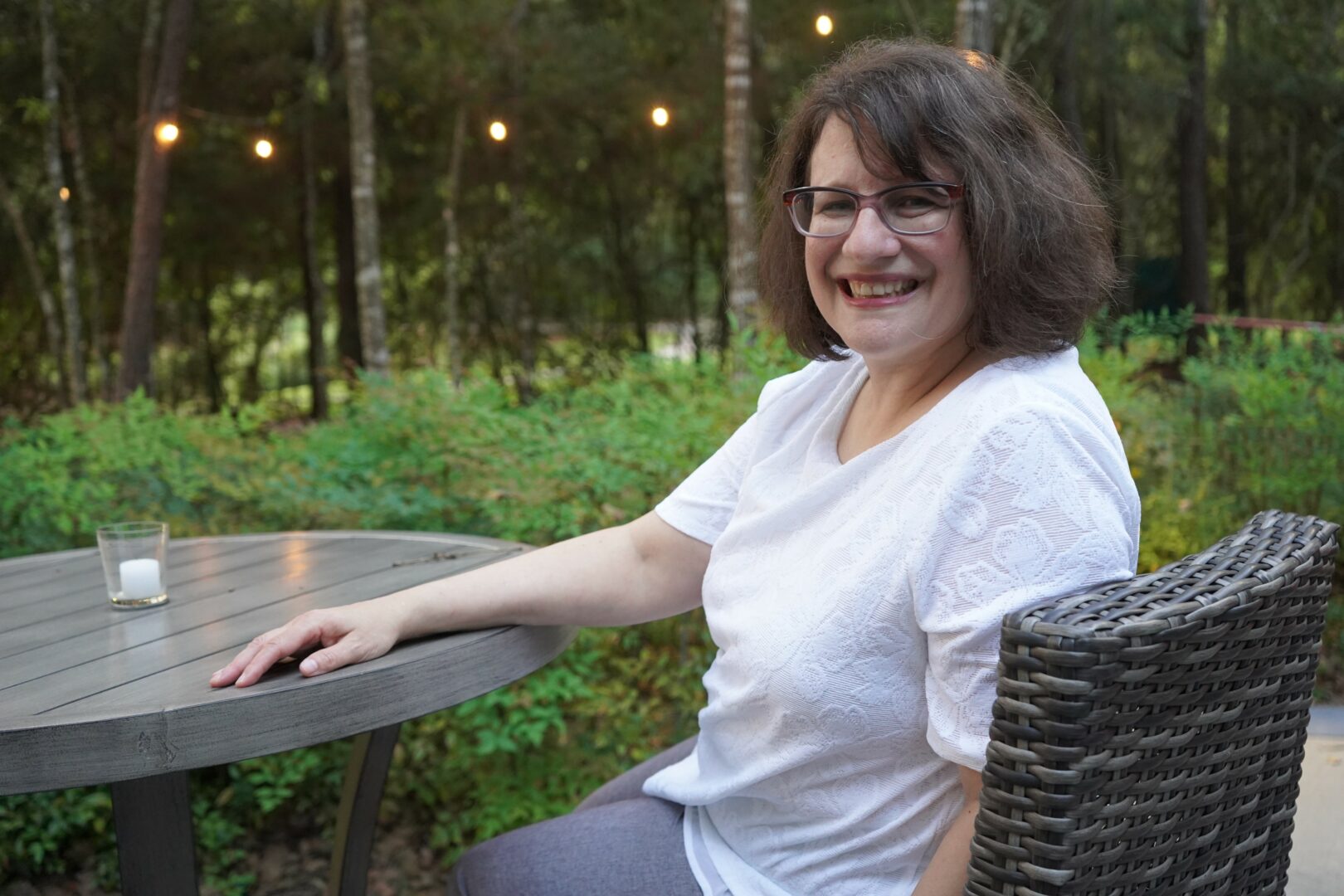 Woman with glasses smiling sitting outdoors at a round table in a wooded area.