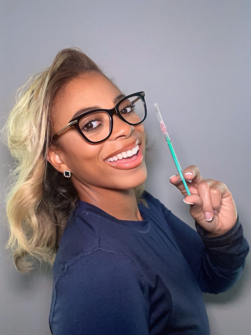 Woman with glasses smiling, holding a syringe, wearing a dark blue shirt, against a plain background.
