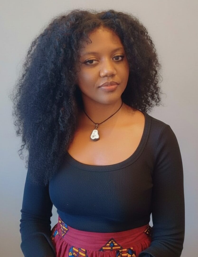 Young woman with curly hair wearing a black top and colorful skirt, standing against a plain background.