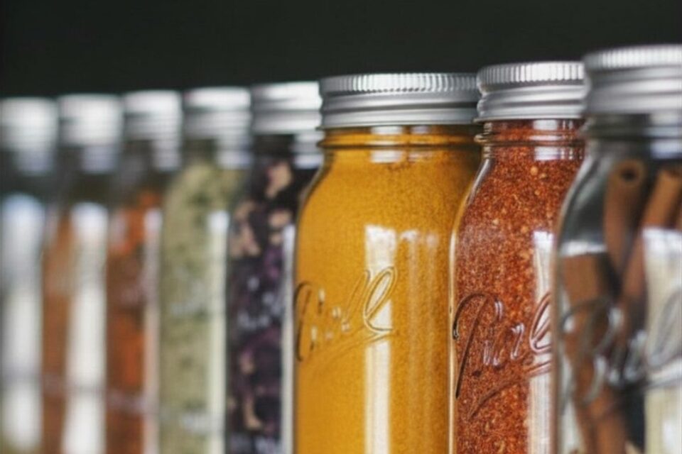 Several glass jars with metal lids containing colorful spices or herbs, arranged in a row on a shelf.
