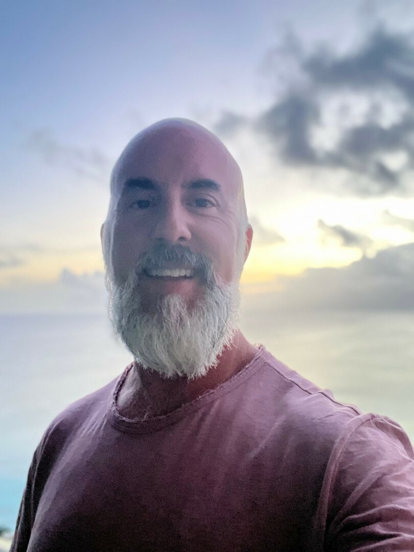 Man with a beard and bald head smiling outdoors with a cloudy sky and ocean in the background.
