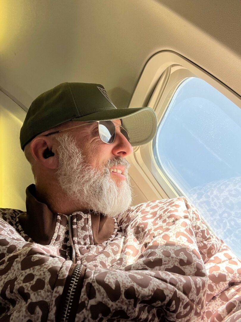 Older man with white beard and glasses, wearing a cap and patterned jacket, resting on an airplane seat with window nearby.