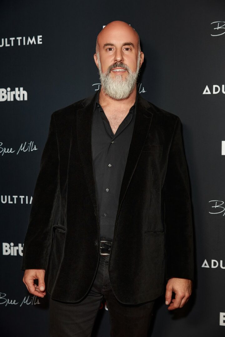 Man with a beard and bald head wearing a black suit and shirt, standing against a dark background with text.