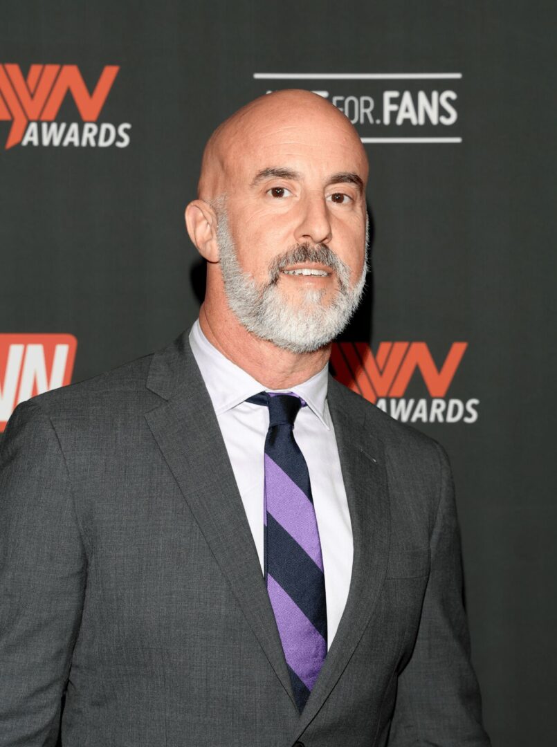 Man with a beard in a suit and tie standing in front of a backdrop with logos.