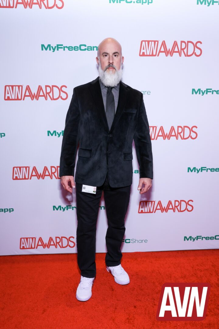 Man with a beard in a dark suit and white sneakers standing on red carpet at awards event.